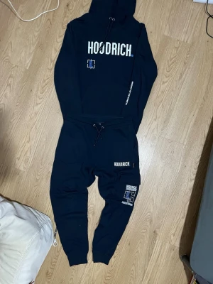 Hoodrich Tracksuit  - Svart tracksuit från Hoodrich med stor vit logga framtill och grafiskt tryck i blått och vitt. Huva med snörning och känguruficka. Materialet är mjuk bomullsmix, perfekt för en avslappnad streetwear-look.