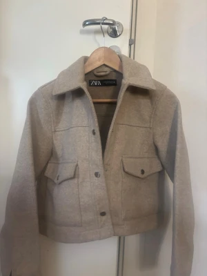 Beige jacka från Zara - Stilren beige jacka  från Zara med klassisk krage, två bröstfickor med lock och knappar, tillverkad i ett mjukt, ull-liknande material som ger en trendig look.