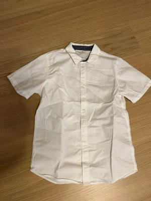 Vit kortärmad skjorta från H&M som ny - Klassisk vit skjorta från H&M med korta ärmar och knappar framtill. Skjortan har en enkel, rak passform och en snygg kontrastkant på insidan av kragen. Perfekt till sommarens alla tillfällen.
