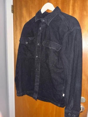 Svart jeansjacka från Jack & Jones - Snygg svart jeansjacka från Jack & Jones med klassisk krage, knappar framtill och två bröstfickor med lock. Tillverkad i ett robust denimtyg som ger en avslappnad och trendig look. Perfekt för lager-på-lager och enkel att styla.