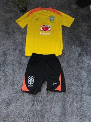 Brasilien fotbollströja och shorts Nike - Gul Brasilien fotbollströja från Nike med gröna och röda detaljer, landslagsemblem och Guaraná-reklam på bröstet. Tillhörande svarta shorts. Perfekt för fotbollsträning eller match. Materialet är lätt och andas bra.