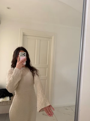 Beige långärmad kort klänning - Trendig beige kort klänning med långärmade, lätt utsvängda ärmar och figurnära passform. Klänningen har en strukturerad yta och rund halsringning. Perfekt för dig som gillar stilrena och enkla plagg med en twist.