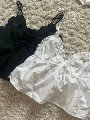 Gina Svart och vit linnetoppar med volang - Två söta linnetoppar i svart och vitt med smala axelband och volangdetaljer längs kanterna. Topparna har en luftig passform och är perfekta för varma dagar. Materialet känns lätt och bekvämt mot huden. 75 kr st 150 för båda
