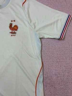 Frankrike fotbollströja Nike vit - Vit fotbollströja från Nike med Frankrikes landslagsemblem och två stjärnor på bröstet. Tröjan har röda och blå detaljer, broderad tupp och FFF-logga, samt randiga muddar i blått, vitt och rött. Perfekt för fotbollsfans som vill visa sitt stöd.