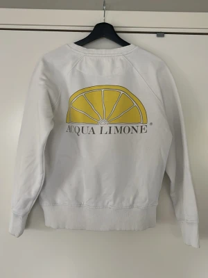 Vit sweatshirt Acqua Limone - Vit sweatshirt från Acqua Limone med klassisk rund hals och tryck både fram och bak. Framsidan har Acqua Limone-loggan och baksidan har en stor gul citronklyfta. Tröjan har ribbade muddar och är tillverkad i mjukt bomullsmaterial. Den har också några flelckar på framsidan med det går säkert att få bort.