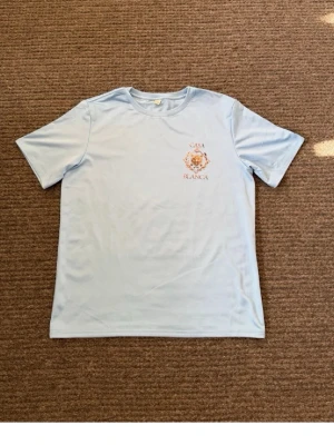 Ljusblå Casa Blanca t-shirt - Snygg ljusblå t-shirt från Casa Blanca med tryck i orange och guld på bröstet och ryggen. Klassisk rund hals och korta ärmar. Perfekt för dig som gillar stilrena plagg med en twist. T-shirten är i mjuk bomull som känns skön mot huden.
