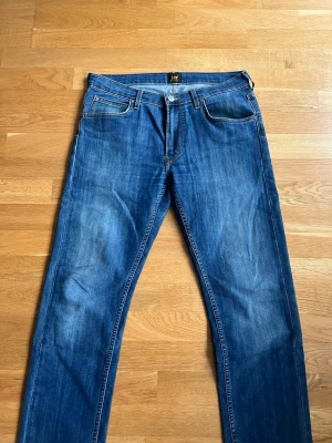 Blå raka jeans från Lee - Snygga blå jeans från Lee med klassisk femficksmodell och raka ben. Jeansen har en mellanblå tvätt med lätt slitning och Lee-logga på bakfickan. Storlek: W32, L32