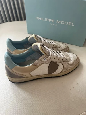 Philippe Model vita och beige sneakers - Snygga sneakers från Philippe Model i vitt och beige läder med ljusblå detaljer på insidan. Skorna har klassisk låg profil, perforerad tå och brunt emblem på sidan. Perfekt för dig som vill ha en stilren och trendig look. I nyskick!! 1500kr. nypris ca 3500kr. Kartong medföljer. Äkta vara!