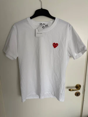 Vit Comme des Garçons Play t-shirt - Vit t-shirt från Comme des Garçons Play med det ikoniska röda hjärtat med ögon broderat på bröstet. Tillverkad i 100% bomull och har en klassisk rund halsringning och korta ärmar. Perfekt för en clean och stilren look.
