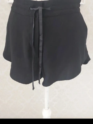 Svarta shorts med snörning - Svarta shorts med elastisk midja och snörning framtill. Modellen är loose fit och har praktiska fickor på sidorna. Perfekta för en avslappnad stil och tillverkade i mjuk polyester inkl foder.