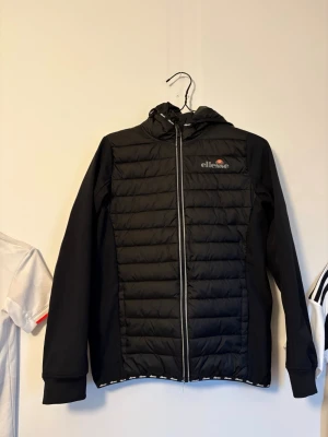 Svart cardiganjacka från Ellesse - Svart pufferjacka från Ellesse med quiltad framsida, huva och hel dragkedja. Jackan har en sportig look med diskret logga på bröstet och släta ärmar. Perfekt för kyliga dagar när du vill vara både varm och snygg.