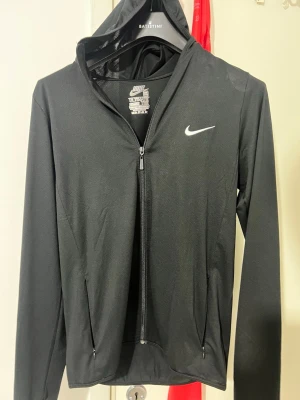 Svart zip hoodie från Nike - Snygg svart hoodie från Nike med dragkedja framtill och vit logga på bröstet. Tillverkad i 100% polyester, perfekt för träning eller chill. Har huva och två fickor med dragkedja. Enkel och sportig design som funkar till det mesta.