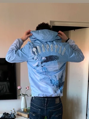 Ljusblå vindjacka från Trapstar - Snygg ljusblå vindjacka från Trapstar med huva och dragkedja. Jackan har stora tryck av örnar på både fram- och baksida samt Trapstar-logga i vitt på ryggen. Två fickor med dragkedja och justerbar nederkant. Perfekt för dig som vill sticka ut med en snygg look och hålla dig varm från vindarna. Tags och påse medföljer!
