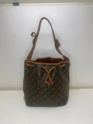 Louis Vuitton Monogram Noé handväska - Klassisk Louis Vuitton Noé handväska i brunt monogrammönstrat canvas med detaljer i läder. Väskan har justerbar axelrem och dragskostängning upptill. Perfekt för dig som gillar tidlös design och vill ha plats för allt du behöver.