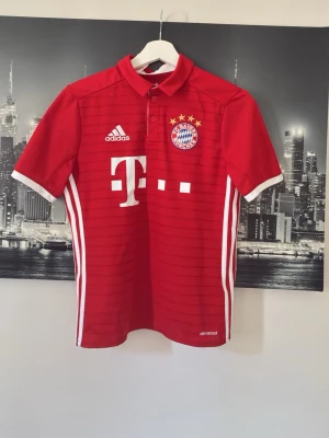 Bayern München matchtröja Adidas - Röd Bayern München matchtröja från Adidas med vita detaljer, klubbmärke och sponsortryck på bröstet samt spelarnamn och nummer 11 (COSTA) på ryggen. Tröjan har korta ärmar, krage med knappar och är tillverkad i funktionsmaterialet climacool passar på en person mellan 160 till 165
