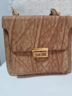 Brun mockaliknande handväska - Säljer en fyrkantig handväska i brun mockaliknande material med unikt mönster. Väskan har guldfärgade detaljer och ett snyggt vridlås framtill. Axelremmen är bred och matchar väskans material. Perfekt accessoar för dig som gillar retrovibbar.