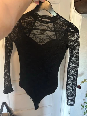 Svart spetsbody med lång ärm - Snygg svart body i spets med långa ärmar och rund hals. Transparent upptill och på ärmarna, med fodrad bystdel för extra täckning. Materialet är stretchigt och följsamt, perfekt för att skapa en trendig look.😍😍