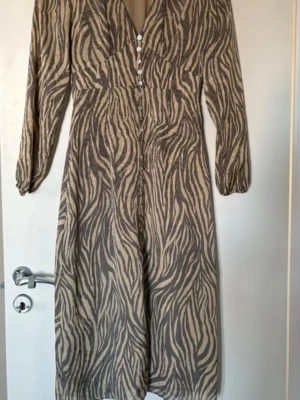 Zebramönstrad långklänning från Adoore - Snygg långklänning från Adoore med zebramönster i beige och grå toner.( ganska liten i storleken)  Klänningen har v-ringning, knappar framtill, långärmade armar och en slits framtill. Perfekt för dig som vill sticka ut med en trendig look.  Klänningen är i modellen med dragkedjan i sidan 💛💛💛