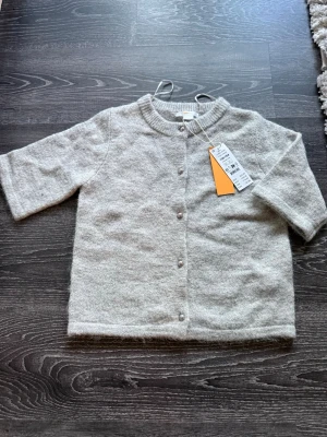 Ljusgrå kofta med kort ärm - Mysig ljusgrå kofta med korta ärmar och rund halsringning. Koftan har knappar framtill och ribbstickad kant vid halsen. Perfekt för lager på lager och enkel att matcha med olika outfits. Från Gina 