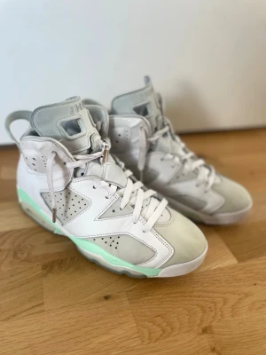 Air Jordan 6 Retro Mint Highs - Säljer ett par Air Jordan 6 Retro sneakers i vitt och ljusgrått med mintgröna detaljer. Skorna har högt skaft, perforerade paneler och klassisk Jumpman-logga på tungan. Materialet är en mix av läder och syntet. Perfekt för dig som vill sticka ut med stilrena sneakers.