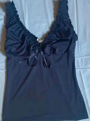 Lila topp  - Snygg svart linnetopp med  v-ringning och volangkant längs axelbanden. Toppen har en liten rosett framtill och är tillverkad i ett mjukt, stretchigt material som känns skönt mot huden. Perfekt för att skapa en chic och enkel look. Jätte fint och i bra skick 🥰