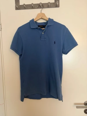 Blå pikétröja från Polo Ralph Lauren - Klassisk blå pikétröja från Polo Ralph Lauren med korta ärmar och krage. Tröjan har två knappar vid halsen och den ikoniska broderade loggan på bröstet. Perfekt till jeans eller shorts för en avslappnad stil.