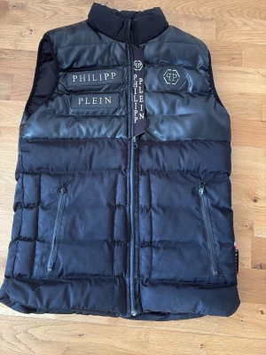 Svart väst från Philipp Plein - Snygg svart dunväst från Philipp Plein med coola detaljer som logotyp på bröstet och märkesnamn på dragkedjan. Västen har två dragkedjeförsedda fickor och en hög krage. Materialet är en mix av dun och syntet, med inslag av skinnimitation upptill.