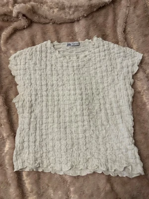 Vit strukturerad topp från Zara - Säljer en vit topp från Zara med strukturerad, våfflad yta. Toppen har rund halsringning och är ärmlös med lätt vågiga kanter. Perfekt för dig som gillar en enkel men ändå unik stil.