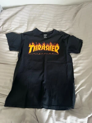 Svart Thrasher t-shirt med flames - Säljer en svart Thrasher t-shirt med den ikoniska loggan i gult och rött flames-tryck på bröstet. Klassisk passform och rund hals. Tillverkad i mjuk bomull, perfekt för en streetwear-look.