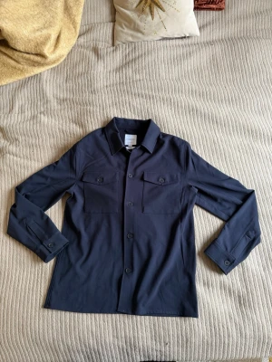 Mörkblå overshirt från Lindbergh - Stilren mörkblå overshirt från Lindbergh med klassisk krage och två bröstfickor med lock och knapp. Rak passform och långa ärmar med knapp vid ärmslut. Perfekt lager-på-lager-plagg i mjukt och lätt material. Jag har använt den fåtal ggr, den är i toppskick och har inga defekter på sig, hör av dig om du har frågor 😁