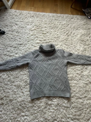 Grå polotröja med stickat mönster - Snygg grå stickad polotröja med stort diamantmönster och ribbade muddar. Tröjan har hög polokrage och är perfekt för kyliga dagar. Materialet känns mjukt och bekvämt, och den har en klassisk passform som funkar till många stilar.