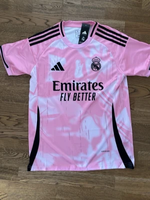 Rosa Real Madrid matchtröja Adidas - Säljer en rosa Real Madrid matchtröja från Adidas med svarta detaljer och klubbmärke på bröstet. Tröjan har svart Adidas-logga, tre svarta ränder på axlarna och trycket 'Emirates Fly Better' på framsidan. Tillverkad i lätt och ventilerande material.