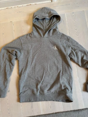 Grå hoodie från Calvin Klein Jeans - Snygg grå hoodie från Calvin Klein Jeans med klassisk huva och diskret 'cK'-logga på bröstet och coolt tryck på ryggen. Tillverkad i mjukt bomullsmaterial som känns skönt mot huden. Perfekt för en avslappnad och stilren look.