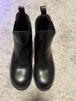 Svarta chelsea boots i skinn - Stilrena svarta chelsea boots i slätt skinn med resår på sidorna och dragflik baktill. Klassisk rund tå och grov sula som ger ett coolt intryck. Perfekta för dig som vill ha en tidlös och trendig look. Ridskor med stålhätta 