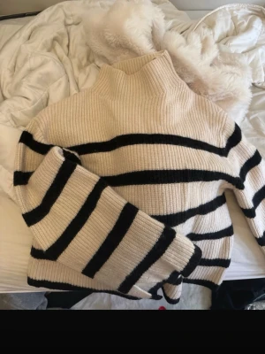 Randig stickad tröja  - Stickad tröja, med breda svarta ränder på beige botten. Tröjan har hög krage och är ribbstickad för extra mysfaktor. Perfekt oversized fit och långa ärmar. Märkt Stockholm Atelier.