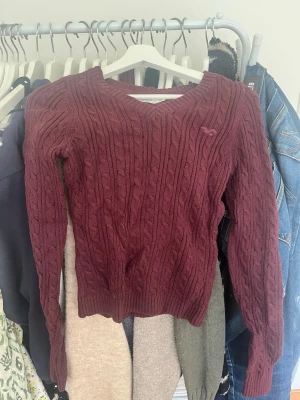 Vinröd kabelstickad tröja V-ringad - Säljer en vinröd kabelstickad tröja med V-ringning och långa ärmar. Tröjan har ett klassiskt kabelmönster och ribbade muddar vid ärmslut och nederkant. Perfekt för dig som gillar en mysig och stilren look.