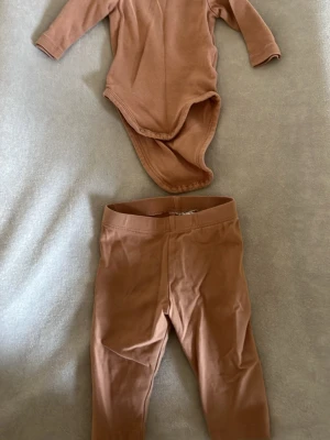 Brunt body- och byxset från H&M - Säljer ett mjukt och bekvämt set från H&M med långärmad body och matchande byxor i en varm brun färg. Perfekt för dig som gillar stilrena och enkla plagg. Materialet är bomull som känns skönt mot huden. Setet har en enkel design utan mönster.