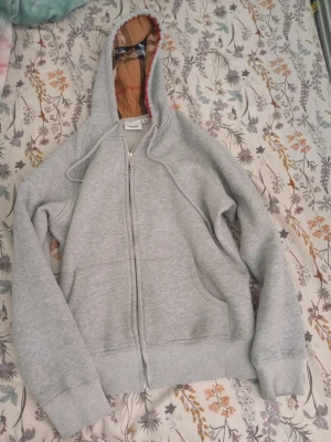 Grå zip-hoodie från Burberry - Säljer en grå zip-hoodie från Burberry med klassiskt rutigt foder i huvan. Hoodien har dragsko, ribbade muddar och två fickor framtill. Materialet är mjukt och bekvämt, perfekt för en chill look. Märket syns tydligt på etiketten i nacken.