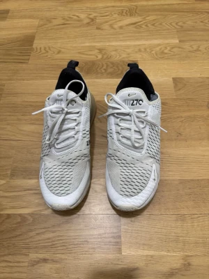 Nike Air Max 270 vita sneakers - Säljer ett par vita Nike Air Max 270 sneakers med mesh-överdel och svarta detaljer. Skorna har en synlig Air-enhet i hälen och klassisk snörning. Lätta och ventilerande med modern design och logga på plösen.