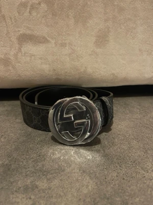 Svart  bälte med silverdetalj - Snyggt svart bälte med klassiskt GG-mönster och stor rund silverfärgad logga som spänne. Bältet har en stilren design och passar perfekt till jeans eller byxor för att lyfta din outfit.
