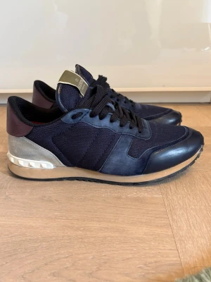 Valentino rockrunners - Snygga sneakers från Valentino Garavani i marinblått tyg och läder med mörkröda detaljer på hälen och grå mocka baktill. Skorna har vita nitar runt hälen och svart sula. Perfekta för dig som vill ha en unik och trendig look. Skorna har en liten defekt se bild 5.