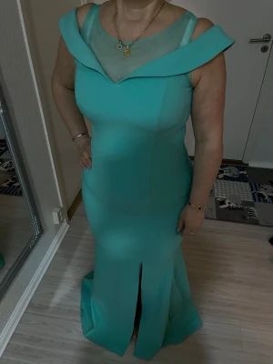 Turkos bröllopsklänning långklänning med meshdetalj - Elegant turkos långklänning med offshoulder-ringning och transparent mesh upptill. Klänningen har en figurnära passform och en slits framtill för extra stil. Perfekt för dig som vill sticka ut med en unik och modern look.