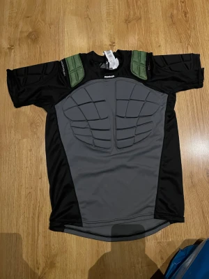 Reebok skyddströja svart/grå - Skyddströja från Reebok i svart och grått med vadderade partier på bröst, rygg och axlar. Kortärmad modell med gröna axeldetaljer. Tillverkad i stretchigt syntetmaterial för bra rörlighet. Barnstorlek