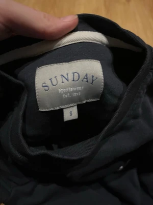 Mörkblå sweatshirt från Sunday - En stilren mörkblå sweatshirt från Sunday Sportswear. Klassisk rund halsringning och långa ärmar. Perfekt för en avslappnad look och enkel att matcha med jeans eller joggers.