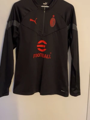 Svart AC Milan träningströja Puma - Svart långärmad träningströja från Puma med AC Milan-logga och röd eFootball-tryck på bröstet. Halvlång dragkedja framtill och gråa detaljer på ärmarna. Tillverkad i ett tekniskt funktionsmaterial som passar perfekt till fotbollsträning.