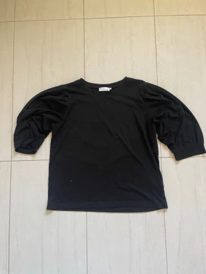 Svart topp med puffärm - Svart topp med rund hals och puffiga trekvartsärmar. Tillverkad i mjukt bomullsmaterial som känns skönt mot huden. Enkel men trendig design som passar till många olika stilar.