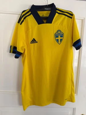 Svensk landslagströja Adidas gul - Gul svensk landslagströja från Adidas med blå detaljer och broderat Sverige-emblem på bröstet. Tröjan har korta ärmar, blå ränder på axlar och ärmslut samt V-ringad krage. Tillverkad i lätt och ventilerande funktionsmaterial.