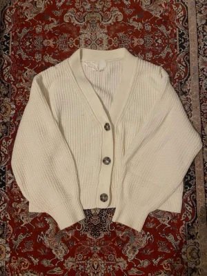 Vit ribbstickad kofta med knappar - Snygg vit ribbstickad kofta med stora silvriga knappar framtill. Koftan har en djup v-ringning, breda muddar och en croppad passform. Perfekt att slänga över en t-shirt eller linne för en chill look.