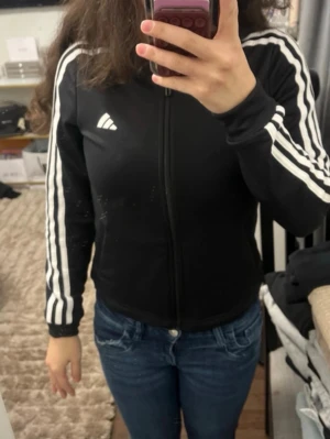 Svart Adidas ziptröja med vita ränder - Säljer en svart ziptröja från Adidas med klassiska vita ränder längs ärmarna och Adidas-logga på bröstet. Tröjan har hög krage och dragkedja framtill. Perfekt till träning eller chill.