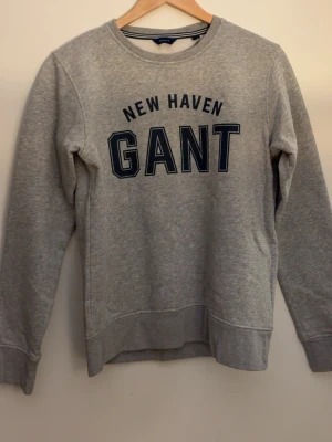 Grå sweatshirt från GANT - Snygg grå sweatshirt från GANT med trycket 'NEW HAVEN GANT' i mörkblå bokstäver på bröstet. Tröjan har rund halsringning, långa ärmar och ribbade muddar vid ärmslut och nederkant. Perfekt för en avslappnad och stilren look.
